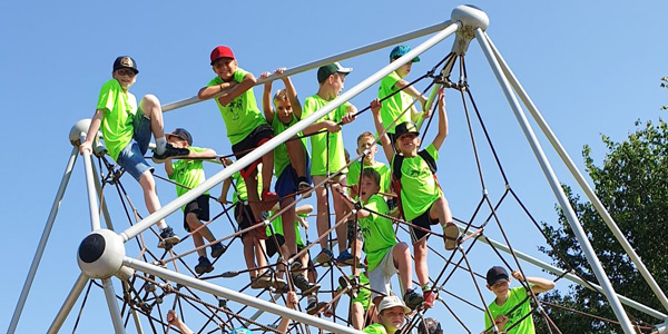 Kinder in neon grünen T-Shirts und Baseballmützen spielen auf einem Seilpyramiden-Klettergerüst vor einem strahlend blauen Himmel.