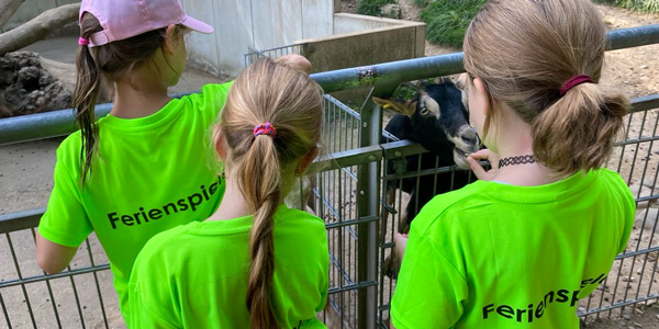 Drei Mädchen mit Pferdeschwanz und neon grünen T-Shirts mit der Aufschrift „Ferienspiele“ stehen vor einem Zaun, hinter dem ein Ziegenbock mit Hörnern zu sehen ist.