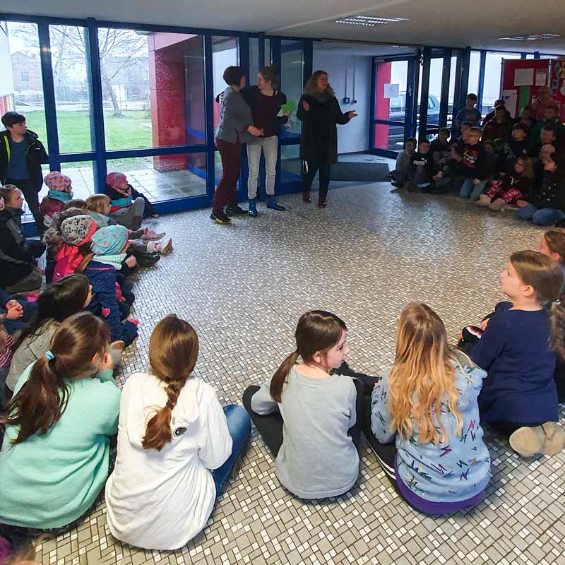 Drei Erwachsene stehen im Foyer vor einer sitzenden Kindermenge.
