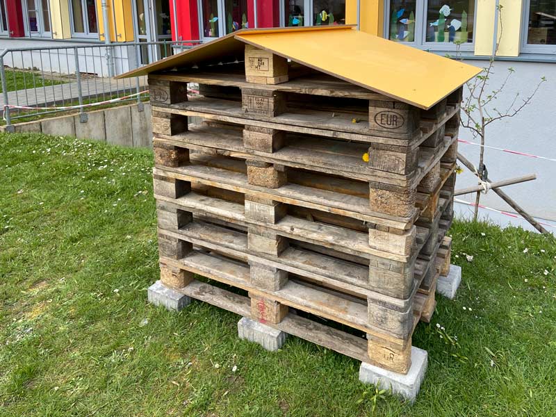 Insektenhotel aus Europaletten auf einer Wiese vor dem Schulgebäude.