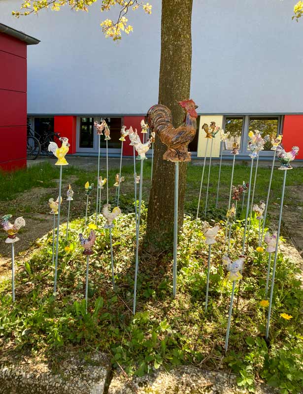Hähne aus Ton, die auf Stangen in ein Beet um einen Baum herum gesteckt wurden.