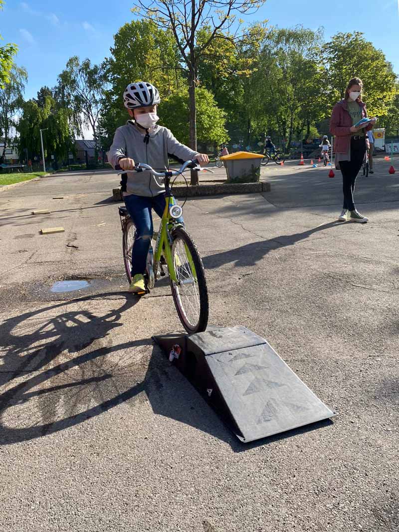 Kind mit Maske und Helm überfährt eine Minirampe mit dem Fahrrad auf dem Schulhof.