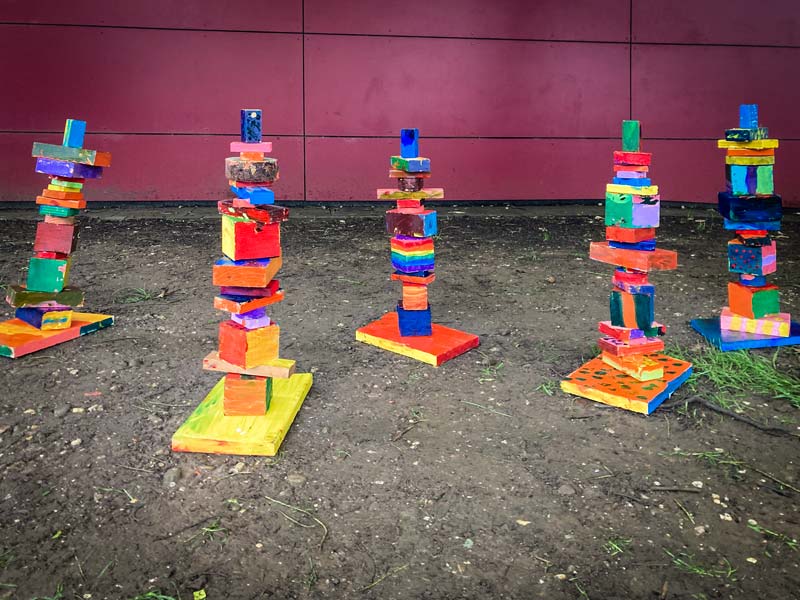 Fünf bunte Holzfiguren stehen auf dem Boden vor einer roten Außenwand.