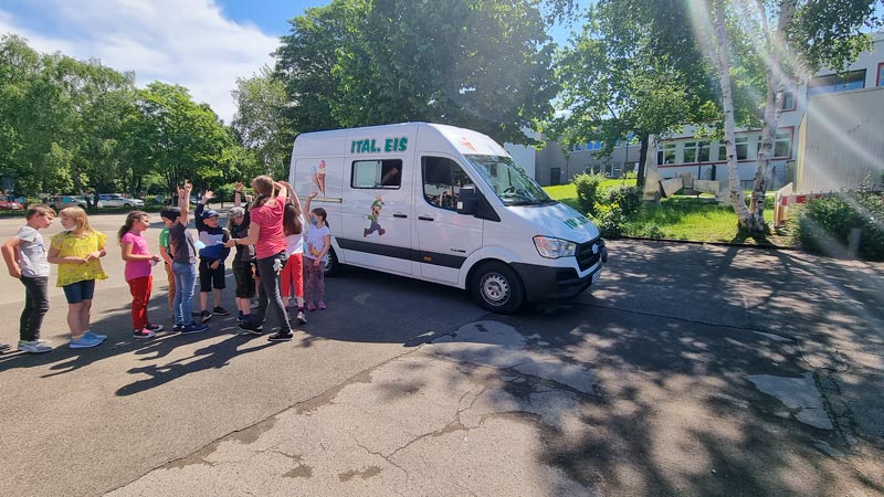 Eine Gruppe von Schulkindern mit Lehrerin stehen in einer Schlange vor einem Eiswagen.
