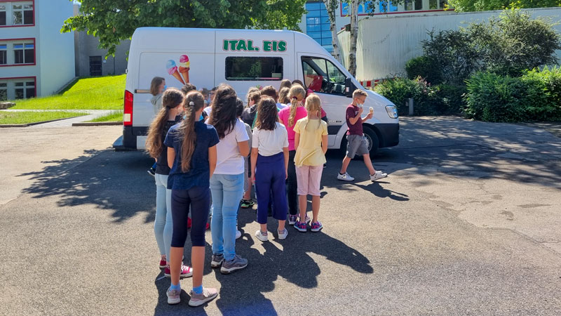 Eine Gruppe von Schulkindern steht auf dem Schulhof in einer Schlange vor einem Eiswagen.