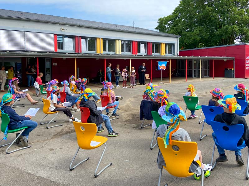 Eine Gruppe von Schulkindern sitzt auf bunten Stühlen auf dem Schulhof.