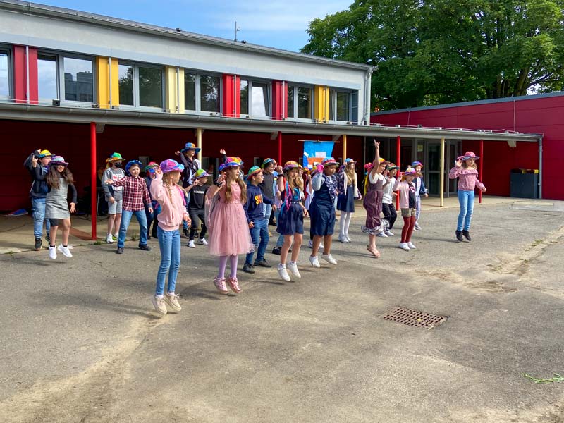 Eine Gruppe von Schulkindern mit bunten Hüten tanzt auf dem Schulhof.