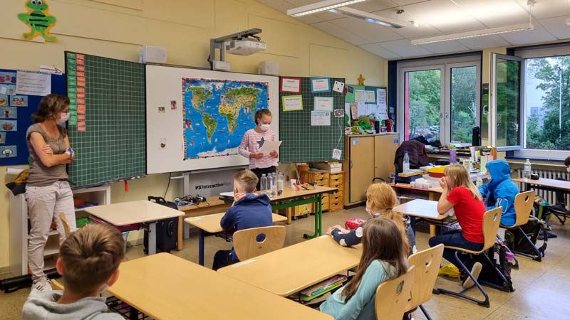 Kinder sitzen im Klassenraum, während eine Schülerin vor der Tafel etwas präsentiert.