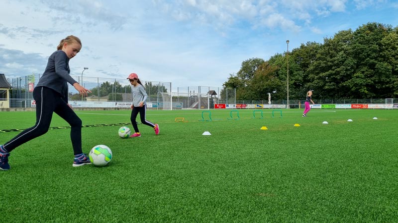 Drei Mädchen laufen Slalom mit einem Ball auf einem Kunstrasenplatz.