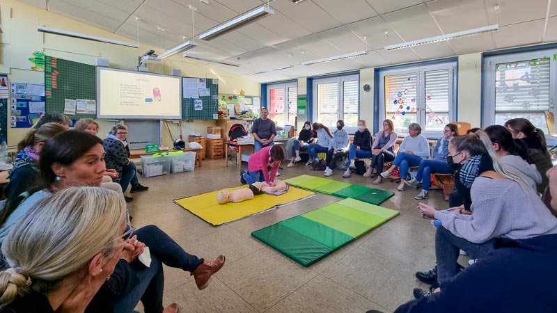 Erwachsene sitzen im Kreis und verfolgen eine Beatmungsübung auf einer Matte im Klassenzimmer.