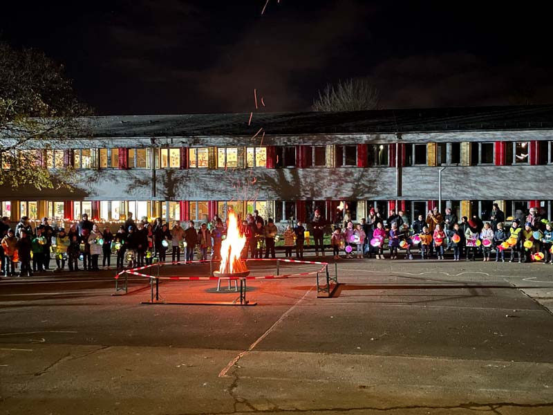 Schulkinder mit Laternen vor dem Schulgebäude stehen am Martinsfeuer.