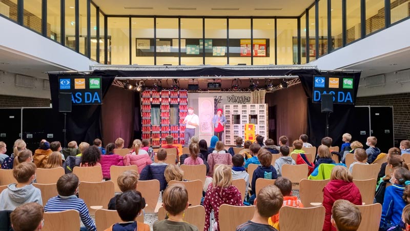 Kinder auf Stühlen schauen auf eine Theaterbühne mit zwei Darstellern.