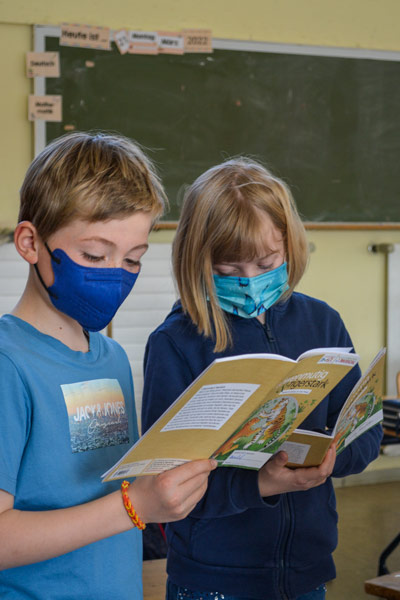 Zwei Kinder mit Maske stehen im Klassenraum und lesen in ihren Heften.