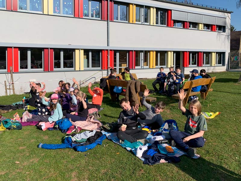 Kindergruppe auf Bänken und Picknickdecken auf dem Rasen vor dem Schulgebäude.