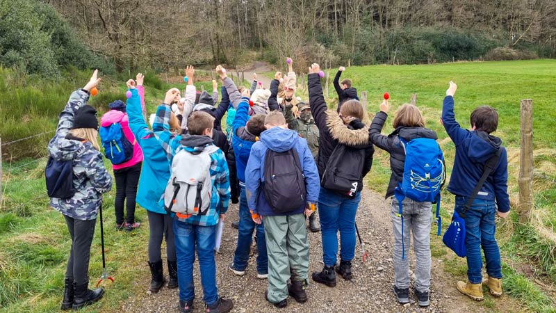 Eine Kindergruppe von hinten auf einem Wanderweg, die alle einen Arm in die Luft strecken und ein Osterei hochhalten.