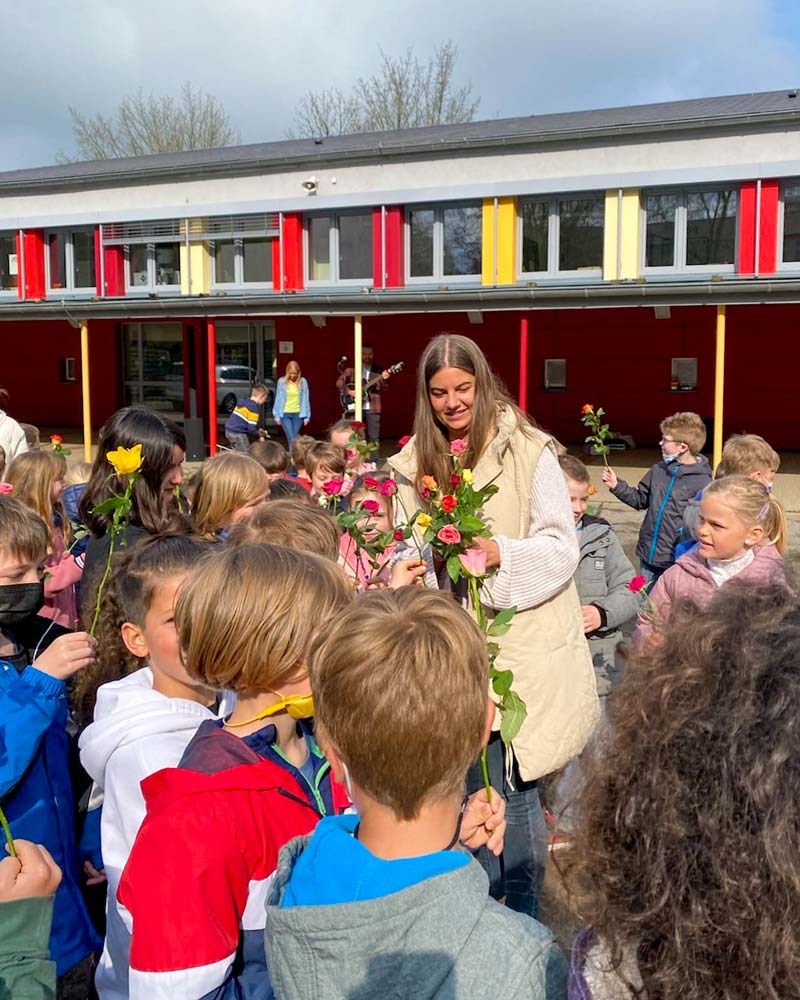 Lehrerin mit Blumen umringt von vielen Kindern auf dem Schulhof.