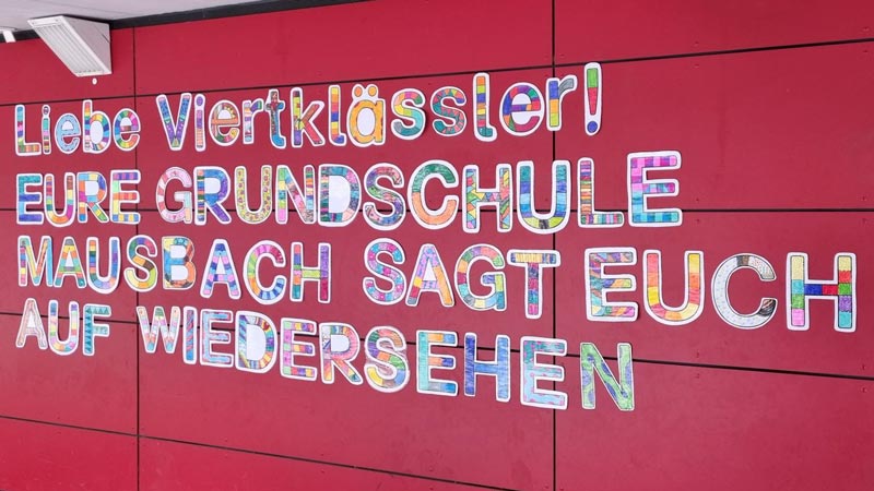 Bunte Buchstaben bilden einen Abschiedsgruß auf einer Wand: Liebe Viertklässler! Eure Grundschule Mausbach sagt euch auf Wiedersehen.