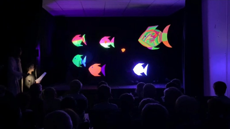 Zuschauer*innen verfolgen im Schwarzlichttheater leuchtende Fische.