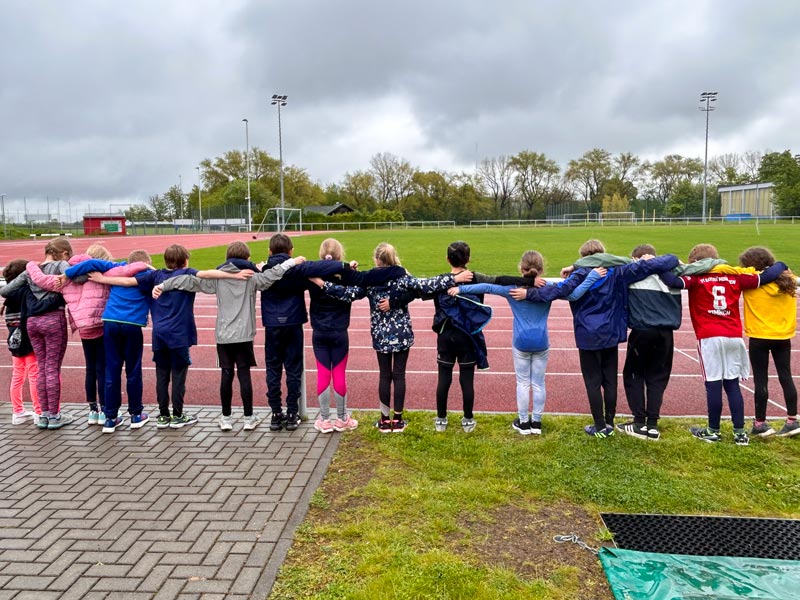 15 Schüler und Schülerinnen von hinten fotografiert umarmt in einer Reihe auf einem Sportplatz.