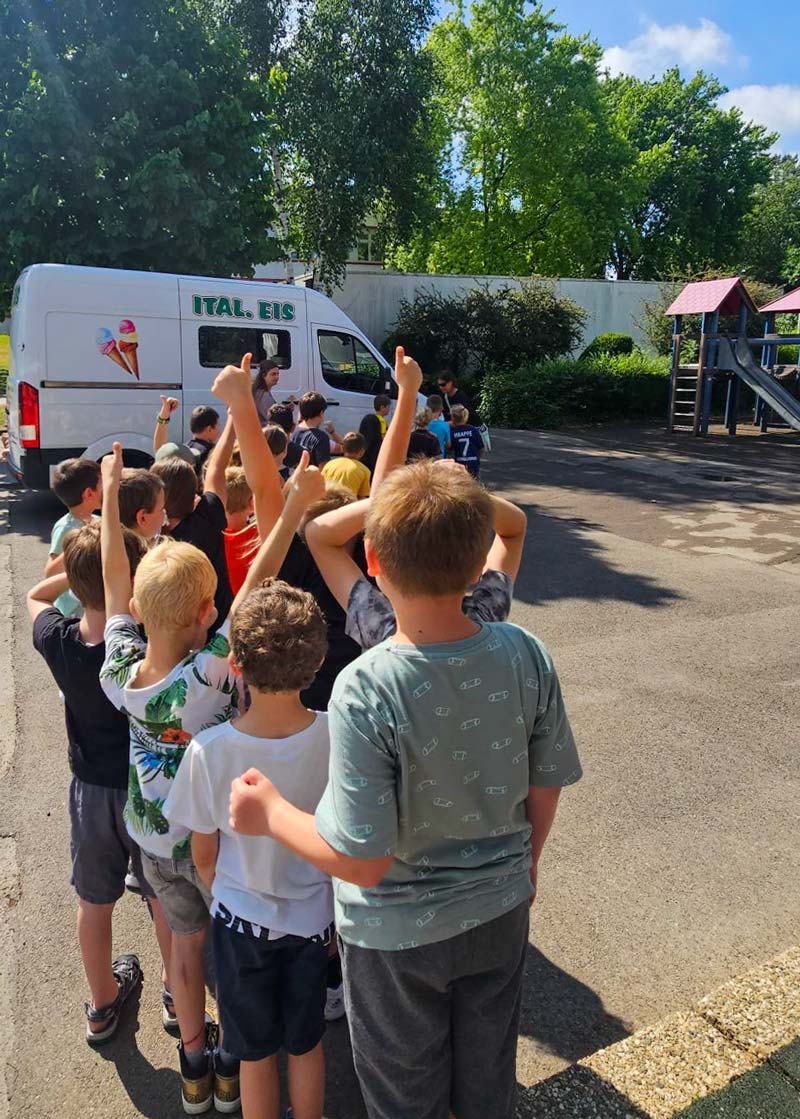 Kinder stehen auf dem Schulhof in einer Reihe vor einem Eiswagen.