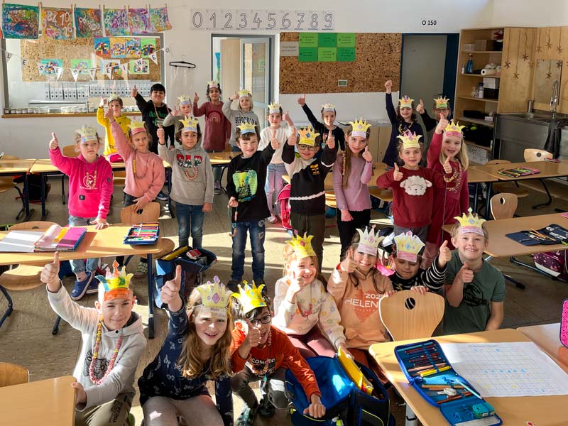 Gruppenfoto der Schulklasse im Klassenzimmer. Die Schüler*innen tragen selbstgebastelte Kronen und strecken einen Daumen in die Luft.