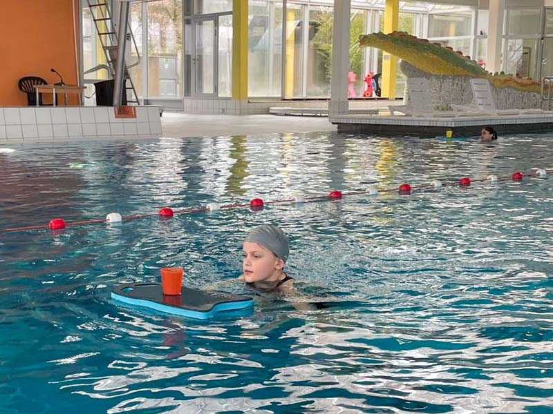 Kind im Wasser einer Schwimmhalle, welches einen Becher auf einem Schwimmbrett vor sich herschiebt.