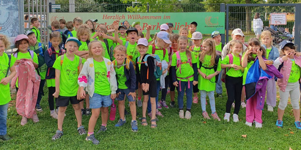 Eine Gruppe von Schulkindern in neongrünen T-Shirts und Rucksäcken posiert vor einem Tor mit einem Schild, auf dem Herzlich Willkommen im Brückenkopf Park steht.