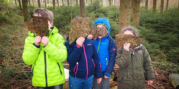 Vier Kinder in bunten Jacken stehen in einem Wald und halten sich jeweils ein Stück Baumrinde vor das Gesicht. Der Hintergrund zeigt einen dichten Wald mit hohen Bäumen und grünem Gestrüpp.
