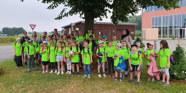 Eine Gruppe von Kindern in passenden neongrünen T-Shirts und mit Rucksäcken, die sich auf einer Wiese mit einem Baum und einem Gebäude im Hintergrund versammelt haben.