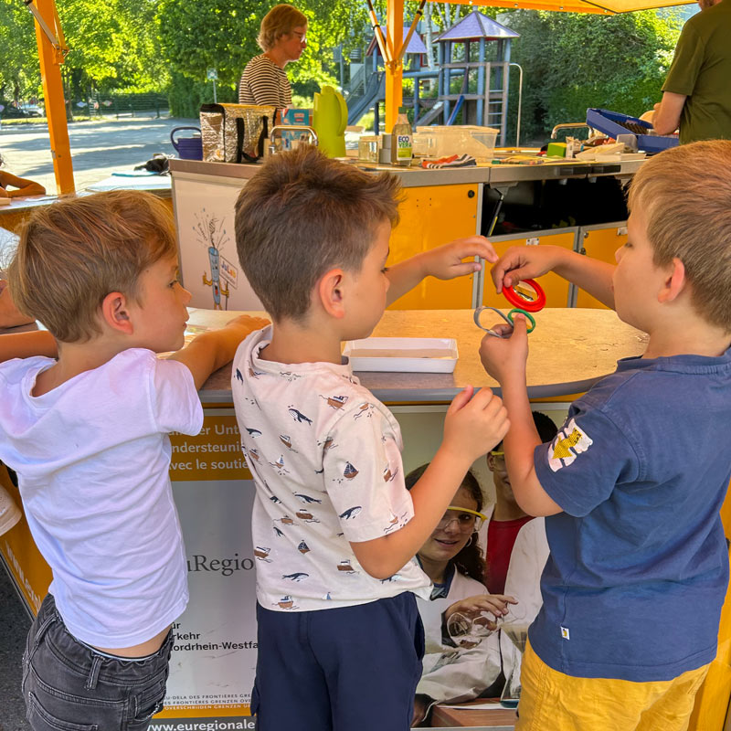 Drei Jungen bei einem Experiment im Freien. Ein Junge schaut zu, während ein anderer bunte Plastikringe hochhält und ein dritter Junge nach ihnen greift. Im Hintergrund ist eine Frau zu sehen.