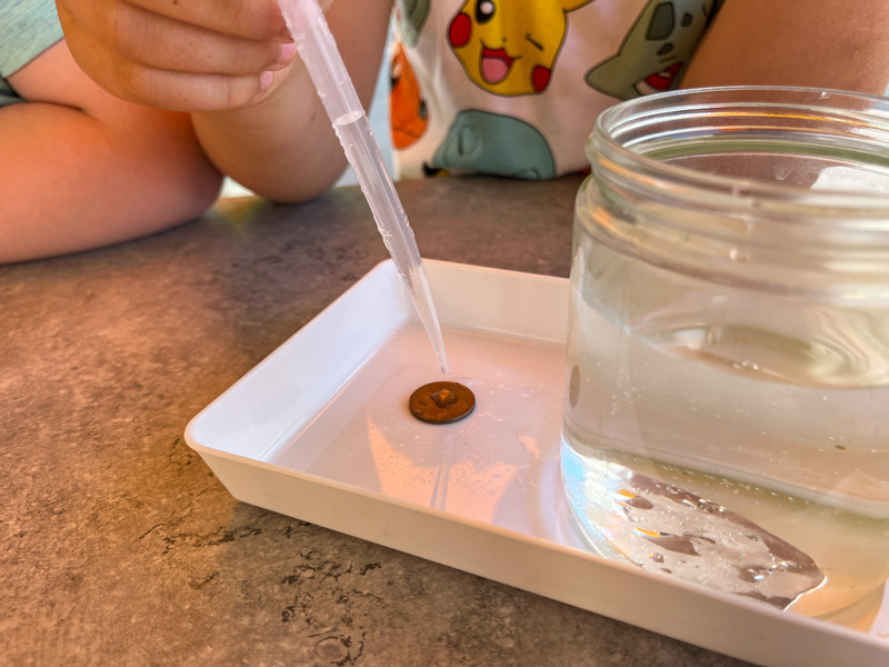 Eine Kinderhand, die mit einer Pipette Wasser auf einen Cent in einem kleinen weißen Tablett tropft, wobei ein Glas mit Wasser zu sehen ist.