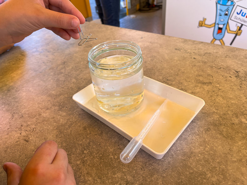 Eine Kinderhand hält eine Büroklammer über einem Glasgefäß mit Wasser, das auf einem weißen Tablett neben einer Pipette steht.