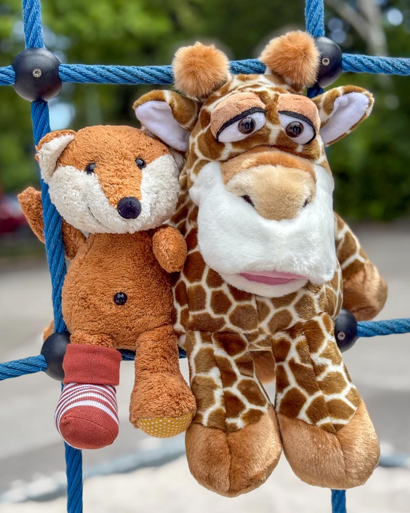 Zwei Plüschtiere, ein Fuchs und eine Giraffe, hängen an einer blauen Seilstruktur vor einem unscharfen Hintergrund im Freien. Der Fuchs ist braun mit einem weißen Gesicht und trägt eine unpassende Socke. Die Giraffe ist braun mit weißen Flecken und einem großen Gesicht.