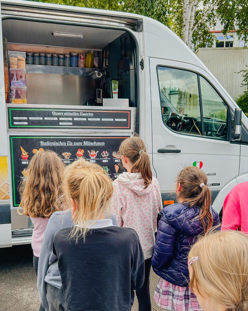 Eine Gruppe junger Mädchen steht in der Schlange vor einem italienischen Eiswagen und betrachtet die Speisekarte, auf der Geschmacksrichtungen wie Vanille, Erdbeere und Kekse angepriesen werden.