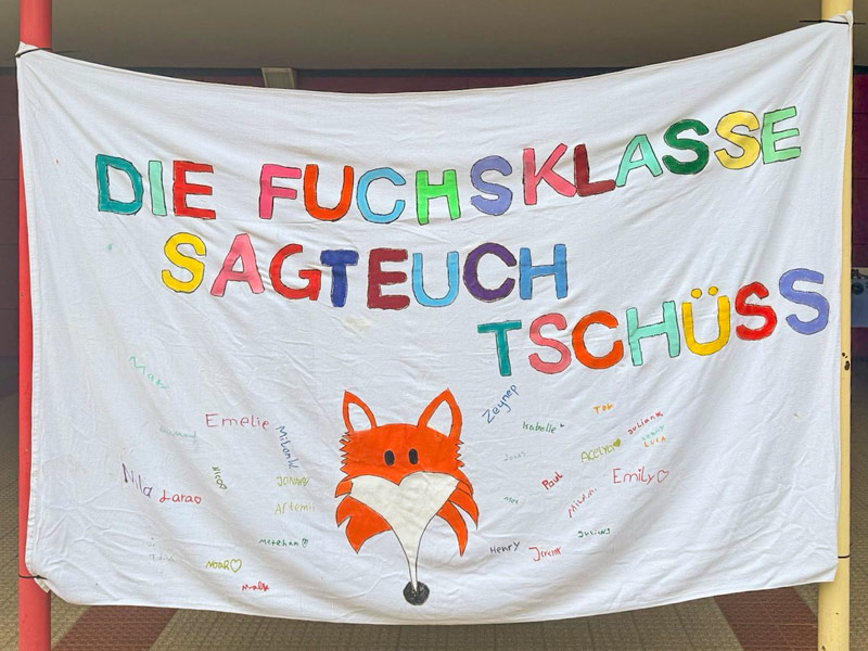 Weißes Banner mit dem Aufdruck DIE FUCHSKLASSE SAGT EUCH TSCHÜSS in bunten Blockbuchstaben, mit einer großen orangefarbenen Fuchs-Illustration in der Mitte und darunter handgeschriebenen Namen.