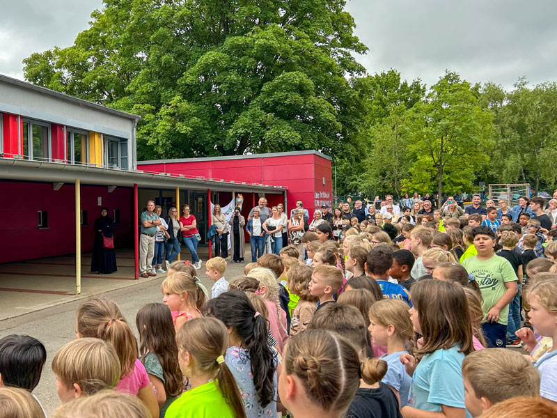 Eine große Gruppe von Kindern steht vor einem Schulgebäude, einige Erwachsene stehen im Hintergrund.