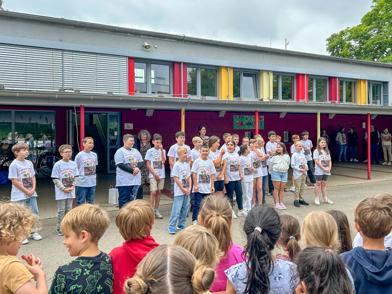 Kinder in weißen T-Shirts mit passendem Aufdruck versammeln sich vor einem Schulgebäude, eine Schar anderer Kinder steht ihnen gegenüber. Auf den T-Shirts steht FUCHSKLASSE 2024-2025.
