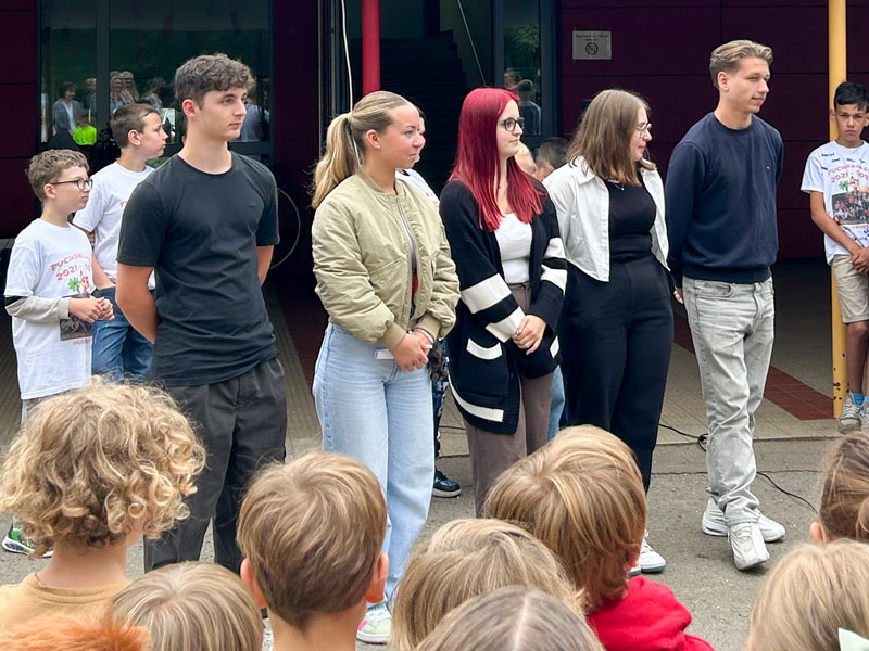 Eine Gruppe von fünf jungen Erwachsenen steht in einer Reihe im Freien, und blickt nach vorne, wobei im Vordergrund mehrere Kinderköpfe zu sehen sind. Im Hintergrund sind einige andere Personen zu sehen.