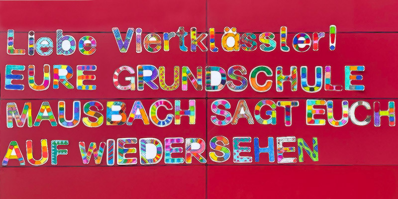 Eine rote Wand mit bunten, ausgeschnittenen Buchstaben mit der Aufschrift LIEBE VIERTKLÄSSLER! EURE GRUNDSCHULE MAUSBACH SAGT EUCH AUF WIEDERSEHEN.