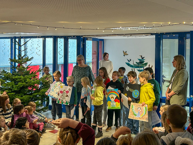 Gruppe von Kindern steht vor einem Weihnachtsbaum und hält bunte Bilder mit Motiven wie Regenbogen, Erde und Engel, mehrere Erwachsene beobachten.