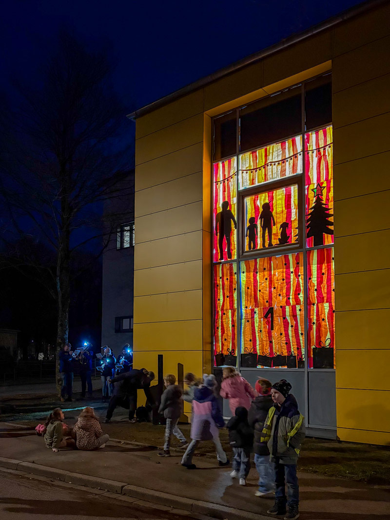 Kinder stehen und sitzen vor einem Gebäude mit einem großen Fenster, das mit bunten, vertikalen Streifen und schwarzen Silhouetten von Menschen und einem Tannenbaum dekoriert ist.