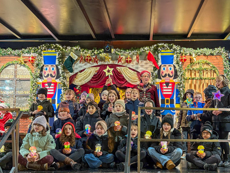 Gruppe von Kindern mit bunten Lichtern in den Händen vor festlich geschmückter Bühne mit Girlanden und Sternen.