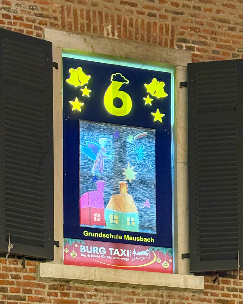 Leuchtendes Adventskalenderfenster mit der Zahl 6, zwei Glocken, mehreren Sternen und einem Bild mit Häusern und einer Figur, darunter der Schriftzug „Grundschule Mausbach“ und Werbung für „BURG TAXI“.