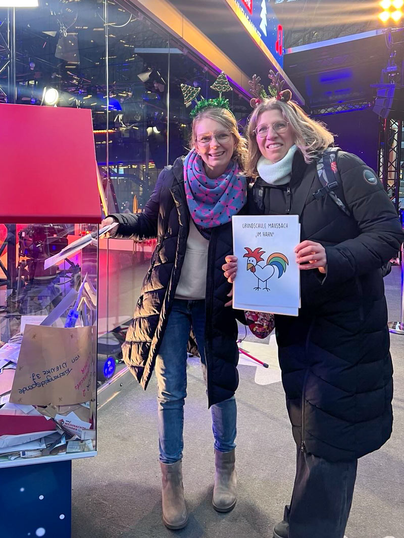 Zwei Frauen mit Winterjacken und Stirnbändern mit Weihnachtsmotiven stehen nebeneinander, eine hält ein Blatt mit einem bunten Hahn und der Aufschrift Grundschule Mausbach „Im Hahn“. Die zweite Frau wirft einen Umschlag in eine große Glasbox.