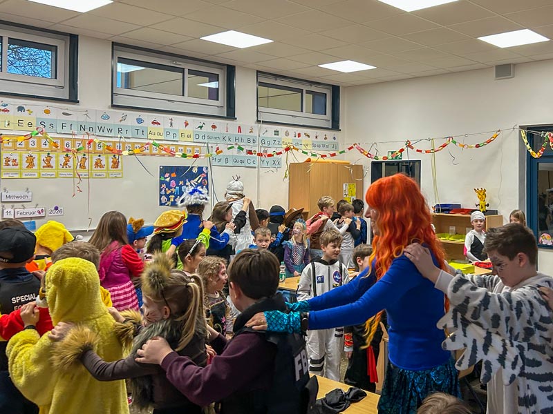 Kinder in einem Klassenzimmer stehen in einer Reihe und halten sich an den Schultern, viele tragen bunte Kostüme und Verkleidungen.