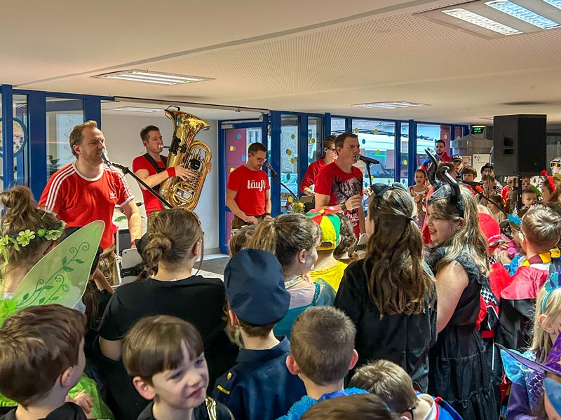 Musiker in roten T-Shirts spielen vor einer Gruppe kostümierter Kinder in einem hellen Raum mit großen Fenstern.