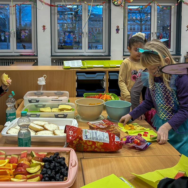 Kind in Meerjungfrauenkostüm nimmt Snacks von einem Tisch mit Obst, Gemüse, Brot und Süßigkeiten in einem Klassenraum mit Fenstern und bunter Dekoration.
