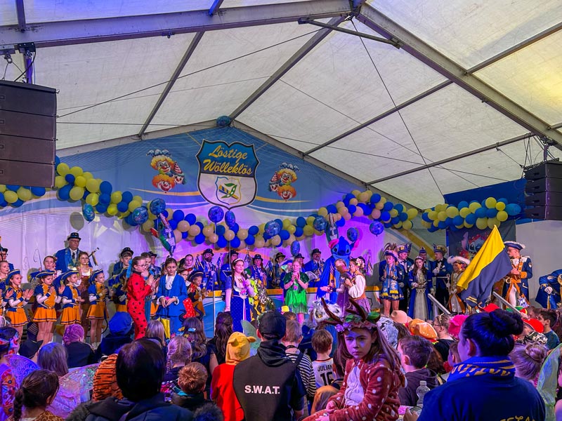 Bühnenauftritt einer bunt kostümierten Gruppe vor Publikum in einem Festzelt mit blau-gelben Luftballons und einem Schild mit der Aufschrift „Löstigen Wölleklös“.