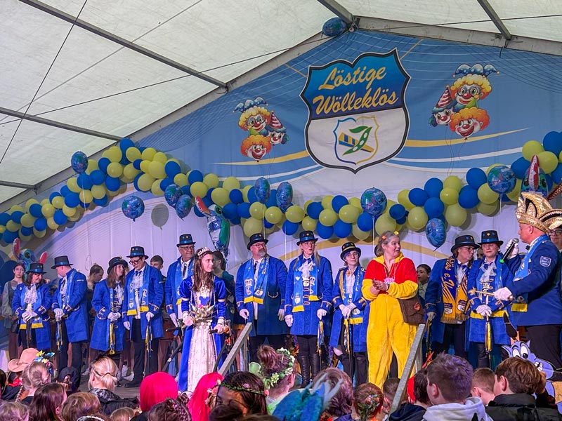 Gruppe von Menschen in blauen Uniformen, mit einer Person in einem gelb-roten Kostüm auf einer Bühne unter einem Banner mit der Aufschrift „Löstigen Wölleklös“ und Clownmotiven, umgeben von blauen und gelben Luftballons.