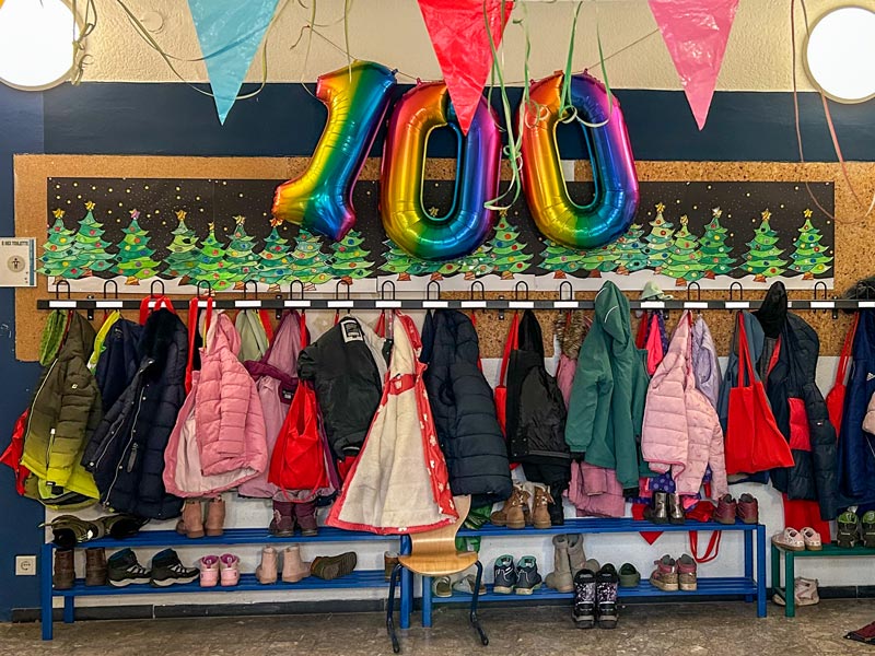 Bunte Luftballons mit der Zahl 100 hängen über einer Garderobe mit Kinderjacken, Schuhen und Taschen vor einer Wand mit Weihnachtsbaum-Malerei.